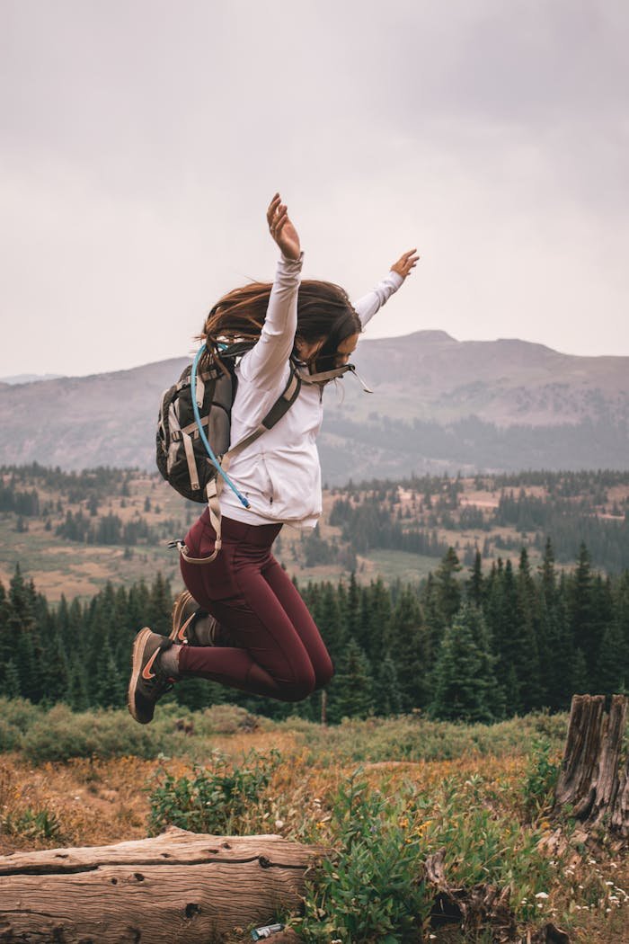 services-img Joyful young woman leaps over a log in a scenic mountainous forest setting, embodying outdoor adventure.